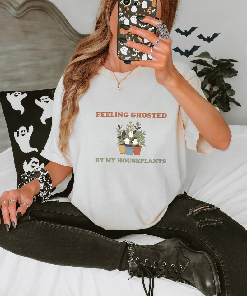 Ivory Comfort Colors t-shirt printed with the words Feeling Ghosted By My Houseplants and a drawing of a compact, self-contained group of three small potted houseplants with tiny, friendly whit, worn by a woman