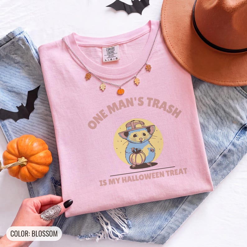 Blossom Comfort Colors t-shirt printed with the words One Man'S Trash Is My Halloween Treat and a drawing of a scruffy opossum wearing a tiny plastic orange pumpkin trick-or-treat bucket on its head, worn by a woman