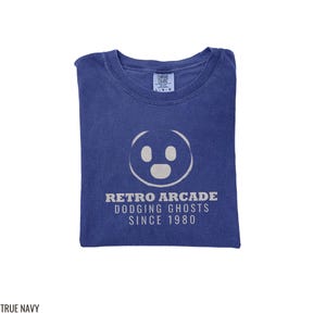 True Navy Comfort Colors t-shirt printed with the words Retro Arcade Dodging Ghosts Since 1980 and a drawing of a maze with a Pac-Man character, folded flat lay