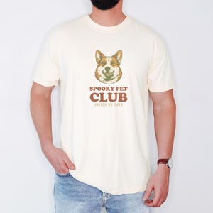 Ivory Comfort Colors t-shirt printed with the words Spooky Pet Club United We Shed and a drawing of a circular crest showing a corgi spirit and a black feline nose-to-nose, worn by a man