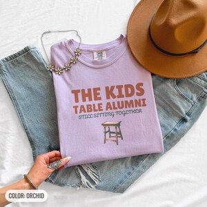 Orchid Comfort Colors t-shirt printed with the words The Kids Table Alumni Still Sitting Together and a drawing of a low wooden folding stool, folded flat lay