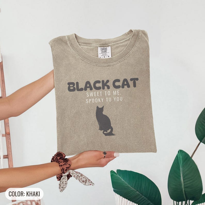 Khaki Comfort Colors t-shirt printed with the words Black Cat Sweet To Me, Spooky To You and a drawing of a fluffy cat that is SOLID JET BLACK from nose to tail, whole body and chest black, sittin, folded flat lay