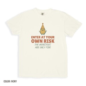 Ivory Comfort Colors t-shirt printed with the words Enter At Your Own Risk The Monsters Are Only Five and a drawing of a cute, smiling little green monster wearing a tiny paper party hat and holding a crayon, folded flat lay