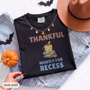 Black Comfort Colors t-shirt printed with the words Thankful Mostly For Recess and a drawing of a stack of retro school books topped with a glossy red apple and a steaming ceramic coffee, worn by a woman