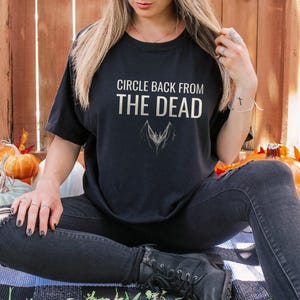 Black Comfort Colors t-shirt printed with the words Circle Back From The Dead and a drawing of a vintage silver paperclip bent into the shape of a bat, worn by a woman