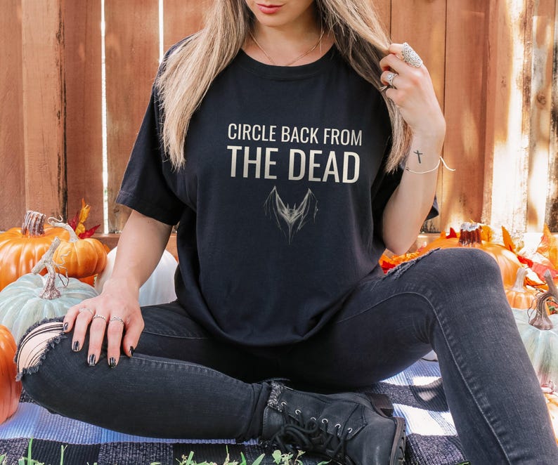 Black Comfort Colors t-shirt printed with the words Circle Back From The Dead and a drawing of a vintage silver paperclip bent into the shape of a bat, worn by a woman
