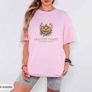 Blossom Comfort Colors t-shirt printed with the words Swapping Spooky For Festive Spirit and a drawing of a spooky jack-o-lantern pumpkin alongside a festive holiday wreath, worn by a woman