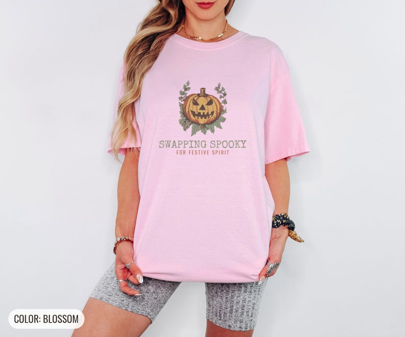 Blossom Comfort Colors t-shirt printed with the words Swapping Spooky For Festive Spirit and a drawing of a spooky jack-o-lantern pumpkin alongside a festive holiday wreath, worn by a woman