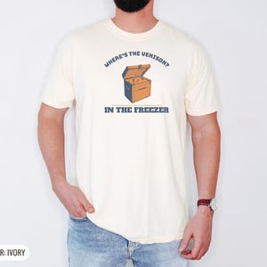 Ivory Comfort Colors t-shirt printed with the words Where'S The Venison? In The Freezer and a drawing of a vintage metal chest freezer with a pair of paper-wrapped butcher packages resting on top, worn by a man