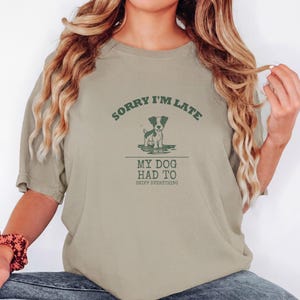Khaki Comfort Colors t-shirt printed with the words Sorry I'M Late My Dog Had To Sniff Everything and a drawing of a brown and white terrier standing in a puddle, worn by a woman