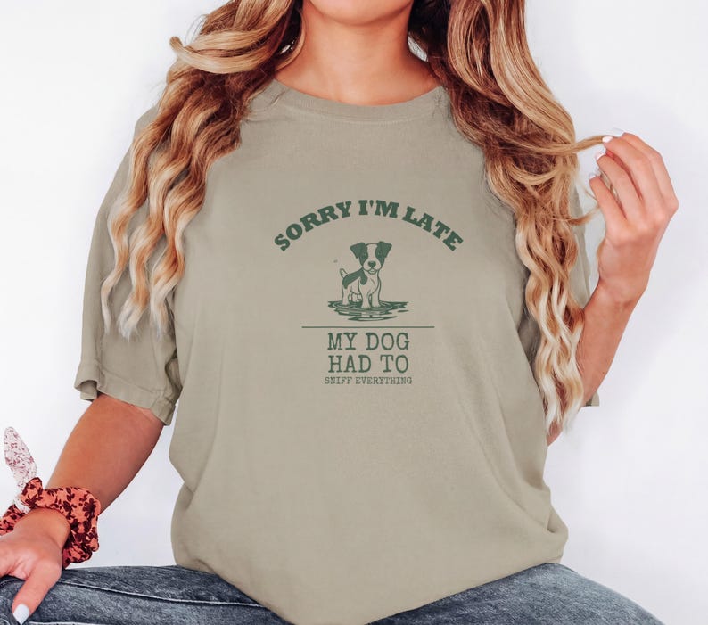 Khaki Comfort Colors t-shirt printed with the words Sorry I'M Late My Dog Had To Sniff Everything and a drawing of a brown and white terrier standing in a puddle, worn by a woman