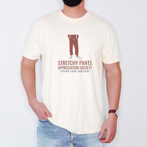 Ivory Comfort Colors t-shirt printed with the words Stretchy Pants Appreciation Society Expand Your Horizons and a drawing of a pair of classic grey sweatpants with a visible drawstring, worn by a man