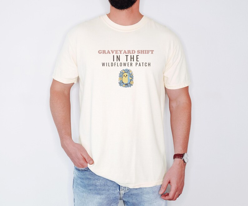 Ivory Comfort Colors t-shirt printed with the words Graveyard Shift In The Wildflower Patch and a drawing of a cute chubby ghost wearing a flower crown and holding a single violet, worn by a man