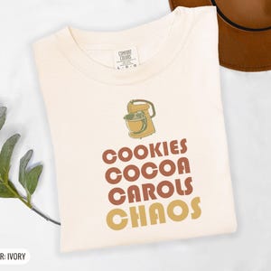 Ivory Comfort Colors t-shirt printed with the words Cookies Cocoa Carols Chaos and a drawing of a retro metal hand mixer with steel beaters, folded flat lay
