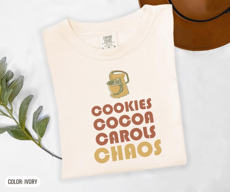 Ivory Comfort Colors t-shirt printed with the words Cookies Cocoa Carols Chaos and a drawing of a retro metal hand mixer with steel beaters, folded flat lay