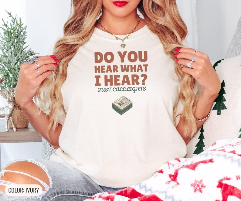 Ivory Comfort Colors t-shirt printed with the words Do You Hear What I Hear? Just Call Lights and a drawing of a rectangular electronic medical pager with a small holly sprig beside it, worn by a woman
