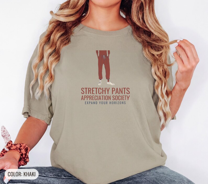 Khaki Comfort Colors t-shirt printed with the words Stretchy Pants Appreciation Society Expand Your Horizons and a drawing of a pair of classic grey sweatpants with a visible drawstring, worn by a woman