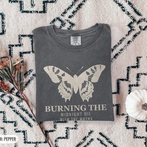 Pepper Comfort Colors t-shirt printed with the words Burning The Midnight Oil With The Moths and a drawing of a cecropia moth with feathery antennae, worn by a woman
