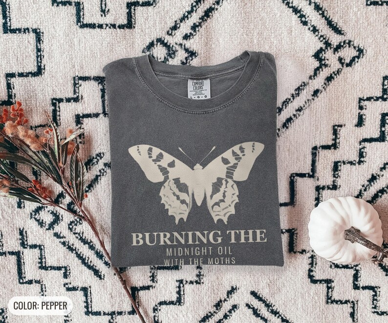 Pepper Comfort Colors t-shirt printed with the words Burning The Midnight Oil With The Moths and a drawing of a cecropia moth with feathery antennae, worn by a woman