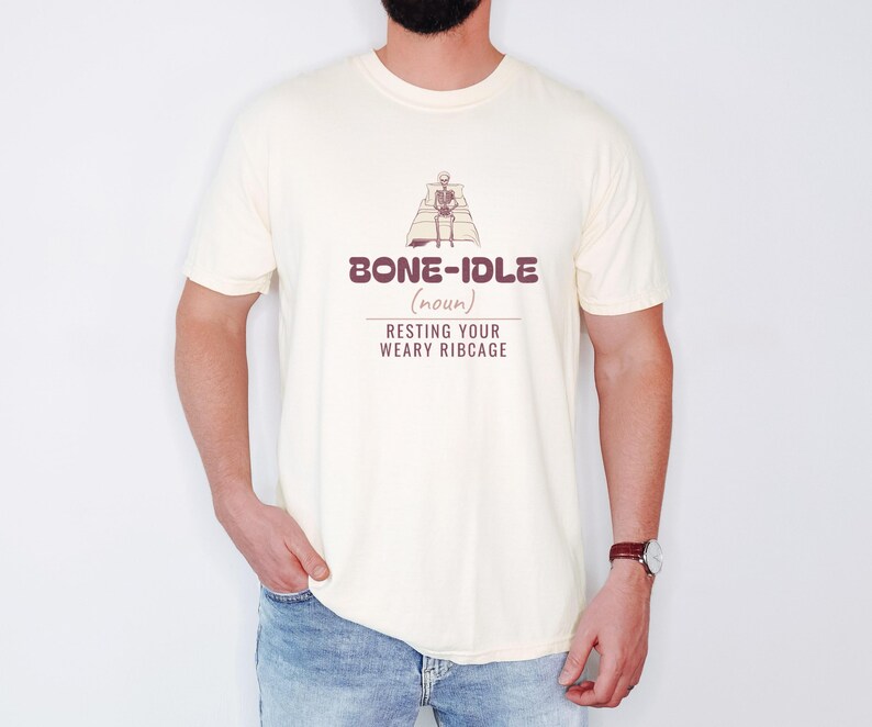 Ivory Comfort Colors t-shirt printed with the words Bone-Idle (Noun) Resting Your Weary Ribcage and a drawing of a skeleton completely tucked under a thick, fluffy duvet in a wooden four-poster bed, worn by a man