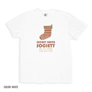 White Comfort Colors t-shirt printed with the words Secret Santa Society We All Know Who Got Who and a drawing of a knitted wool stocking stuffed with stick candy, folded flat lay