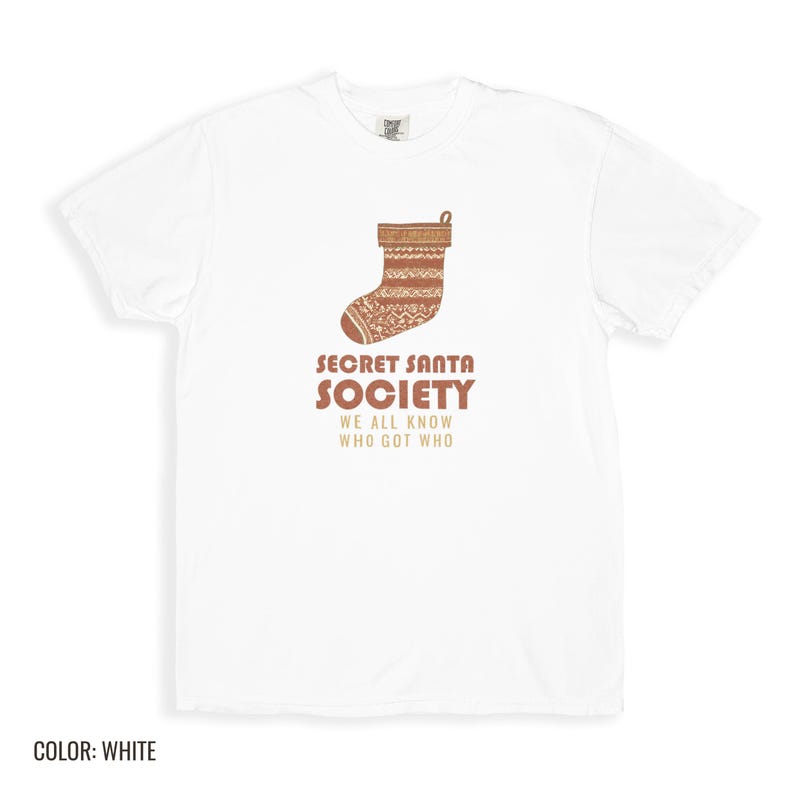 White Comfort Colors t-shirt printed with the words Secret Santa Society We All Know Who Got Who and a drawing of a knitted wool stocking stuffed with stick candy, folded flat lay