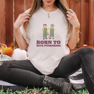 Ivory Comfort Colors t-shirt printed with the words Born To Bite Pumpkins and a drawing of pair of corduroy toddler overalls splattered with pumpkin pulp, worn by a woman