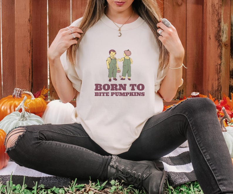 Ivory Comfort Colors t-shirt printed with the words Born To Bite Pumpkins and a drawing of pair of corduroy toddler overalls splattered with pumpkin pulp, worn by a woman