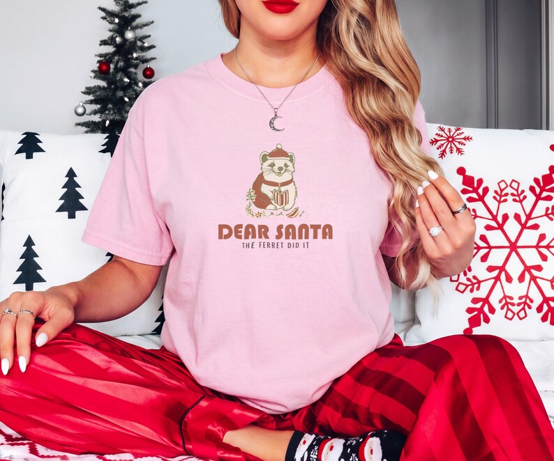 Blossom Comfort Colors t-shirt printed with the words Dear Santa The Ferret Did It and a drawing of a mischievous ferret dragging a striped Christmas stocking filled with shiny ornaments, worn by a woman