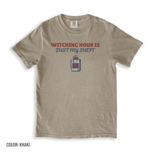 Khaki Comfort Colors t-shirt printed with the words Witching Hour Is Just My Shift and a drawing of a retro medical defibrillator machine decorated with a spooky jack-o-lantern face on its m, folded flat lay