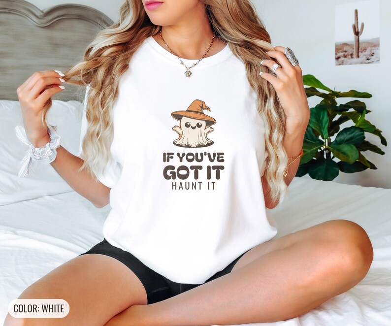 White Comfort Colors t-shirt printed with the words If You'Ve Got It Haunt It and a drawing of a cute retro cartoon ghost wearing a witch hat, worn by a woman