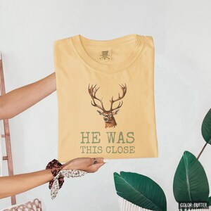 Butter Comfort Colors t-shirt printed with the words He Was This Close and a drawing of a single large twelve-point deer antler shed resting on dry oak leaves, folded flat lay