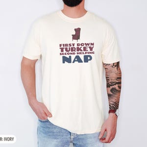 Ivory Comfort Colors t-shirt printed with the words First Down Turkey Second Helping Nap and a drawing of a vintage brown recliner with a footrest extended outward, worn by a man