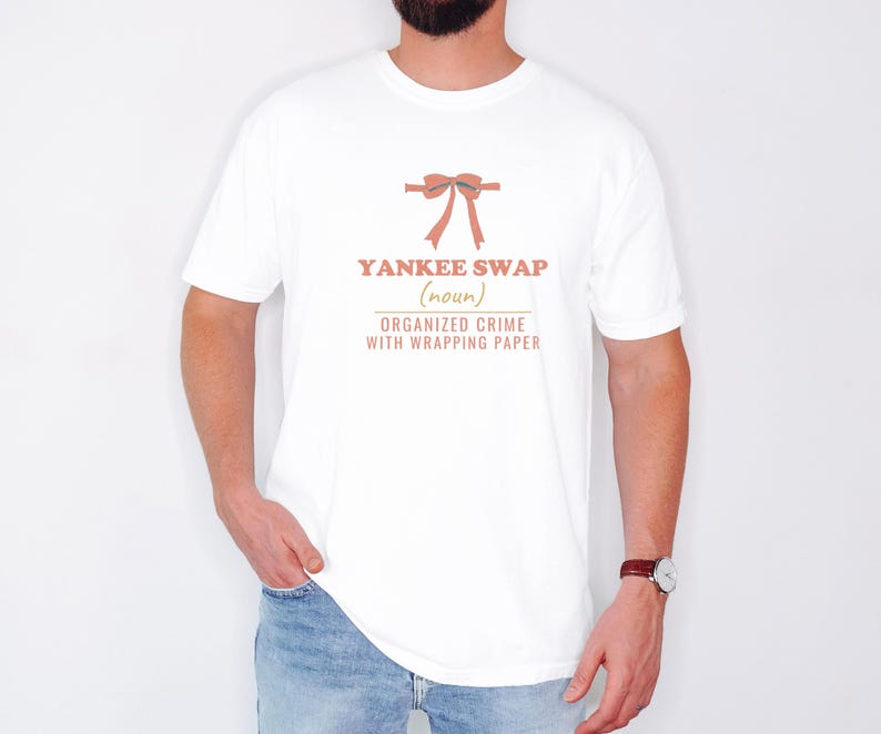 White Comfort Colors t-shirt printed with the words Yankee Swap (Noun) Organized Crime With Wrapping Paper and a drawing of a large red comical bow with long satin ribbon tails, worn by a man