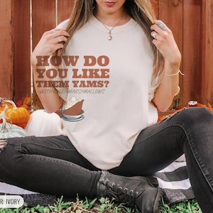 Ivory Comfort Colors t-shirt printed with the words How Do You Like Them Yams? With Extra Marshmallows and a drawing of a roasted yam sliced in half with melted marshmallow topping, worn by a woman