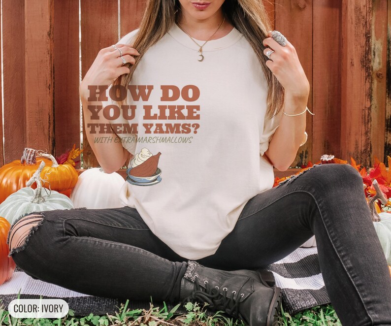 Ivory Comfort Colors t-shirt printed with the words How Do You Like Them Yams? With Extra Marshmallows and a drawing of a roasted yam sliced in half with melted marshmallow topping, worn by a woman