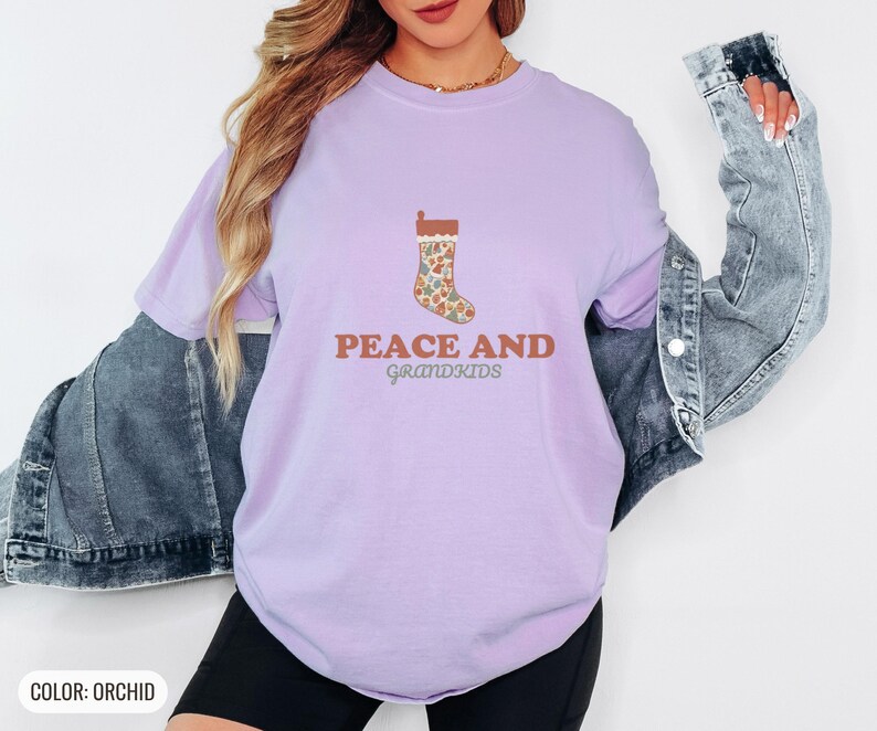 Orchid Comfort Colors t-shirt printed with the words Peace And Grandkids and a drawing of a classic knitted wool christmas stocking stuffed with small toys, worn by a woman