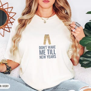 Ivory Comfort Colors t-shirt printed with the words Don'T Wake Me Till New Years and a drawing of a pair of plaid pajama pants folded neatly on a wooden stool, worn by a woman