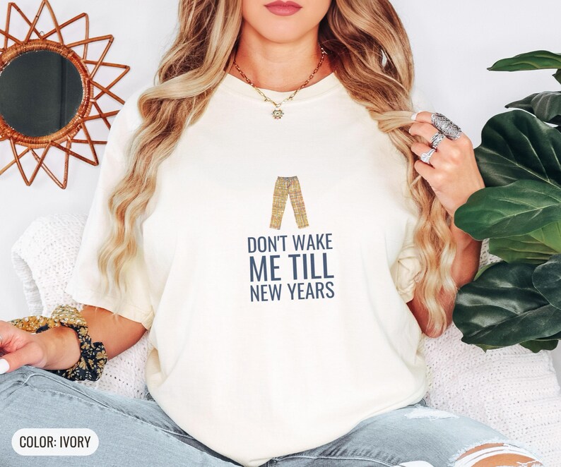 Ivory Comfort Colors t-shirt printed with the words Don'T Wake Me Till New Years and a drawing of a pair of plaid pajama pants folded neatly on a wooden stool, worn by a woman