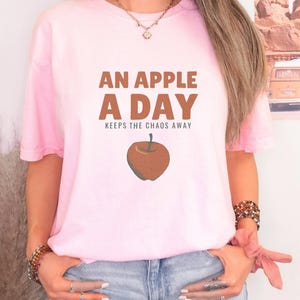Blossom Comfort Colors t-shirt printed with the words An Apple A Day Keeps The Chaos Away and a drawing of a shiny red apple with a single green leaf and a tiny worm peeking out, worn by a woman
