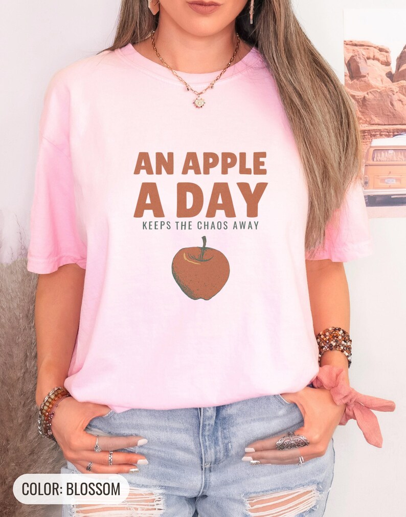 Blossom Comfort Colors t-shirt printed with the words An Apple A Day Keeps The Chaos Away and a drawing of a shiny red apple with a single green leaf and a tiny worm peeking out, worn by a woman