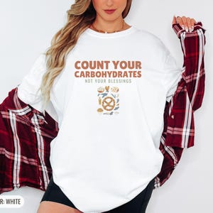 White Comfort Colors t-shirt printed with the words Count Your Carbohydrates Not Your Blessings and a drawing of a golden-brown braided pretzel with coarse salt grains, worn by a woman