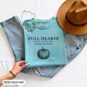 Chalky Mint Comfort Colors t-shirt printed with the words Full Hearts Heavy Plates And Second Helpings and a drawing of a pumpkin surrounded by fall leaves and small acorns, folded flat lay