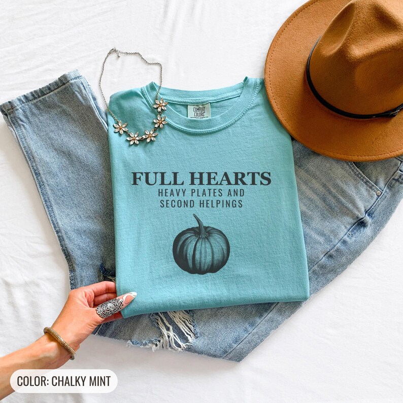 Chalky Mint Comfort Colors t-shirt printed with the words Full Hearts Heavy Plates And Second Helpings and a drawing of a pumpkin surrounded by fall leaves and small acorns, folded flat lay