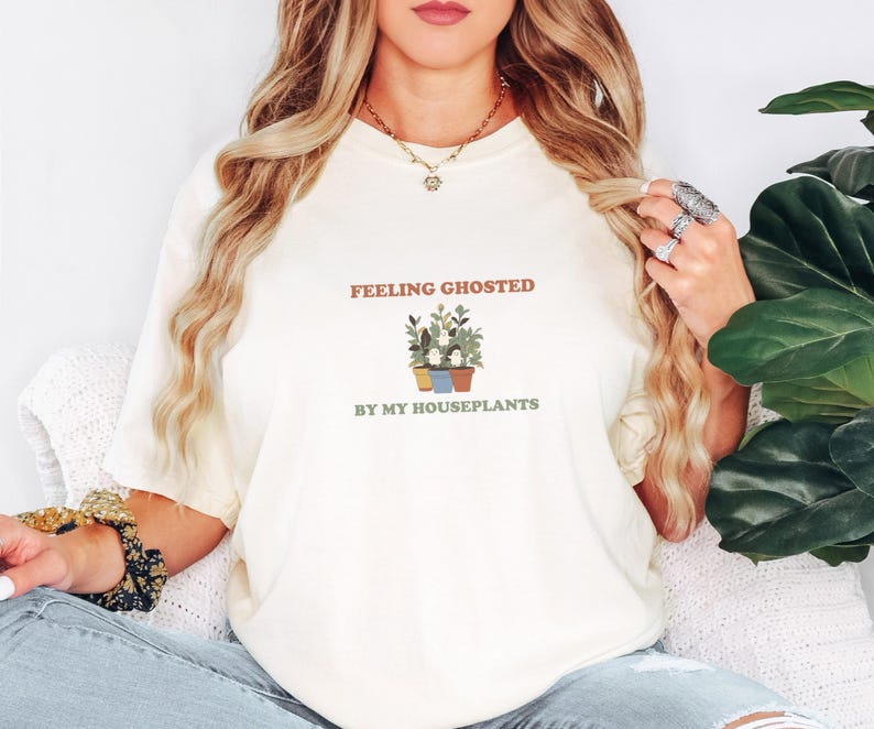 Ivory Comfort Colors t-shirt printed with the words Feeling Ghosted By My Houseplants and a drawing of a compact, self-contained group of three small potted houseplants with tiny, friendly whit, worn by a woman