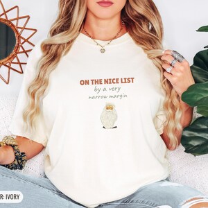 Ivory Comfort Colors t-shirt printed with the words On The Nice List By A Very Narrow Margin and a drawing of a red santa hat beside a candy cane, worn by a woman