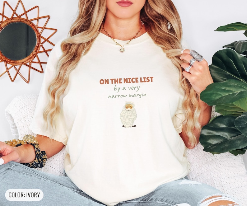 Ivory Comfort Colors t-shirt printed with the words On The Nice List By A Very Narrow Margin and a drawing of a red santa hat beside a candy cane, worn by a woman