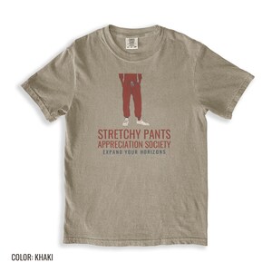 Khaki Comfort Colors t-shirt printed with the words Stretchy Pants Appreciation Society Expand Your Horizons and a drawing of a pair of classic grey sweatpants with a visible drawstring, folded flat lay