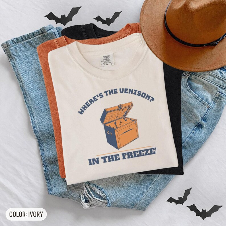 Ivory Comfort Colors t-shirt printed with the words Where'S The Venison? In The Freezer and a drawing of a vintage metal chest freezer with a pair of paper-wrapped butcher packages resting on top, worn by a woman