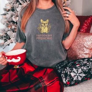 Pepper Comfort Colors t-shirt printed with the words Pardon My Frenchie Under The Mistletoe and a drawing of a vintage brass French Bulldog bottle opener lying next to a sprig of red-berried mistleto, worn by a woman